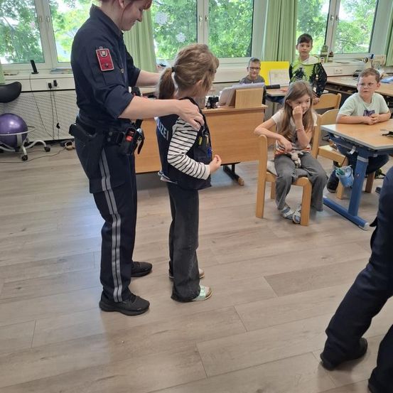 Ein Polizist hilft einem jungen Mädchen im Klassenzimmer. Andere Kinder sitzen an Tischen, und ein Junge beobachtet.