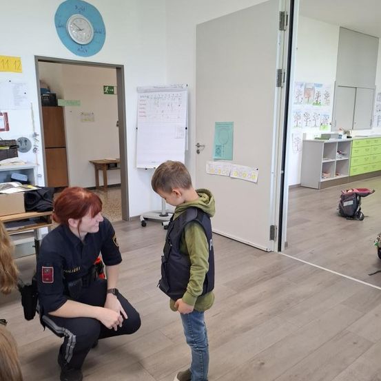 Eine Polizeibeamtin kniet und spricht mit einem Jungen in einem Klassenzimmer. Das Klassenzimmer hat einen Holzboden und eine Tafel mit einem Kalender.