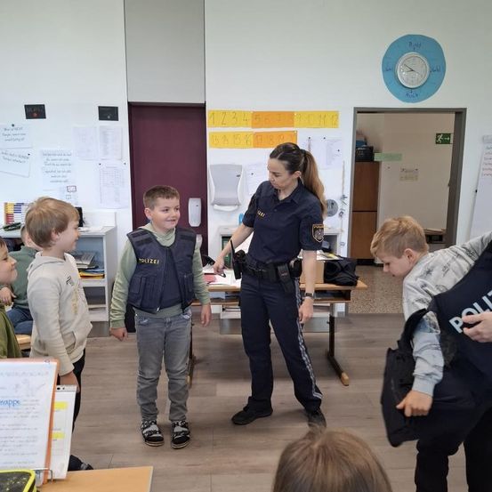 Eine Polizistin in Uniform spricht mit Kindern in einem Klassenzimmer. Die Kinder stehen und lächeln.