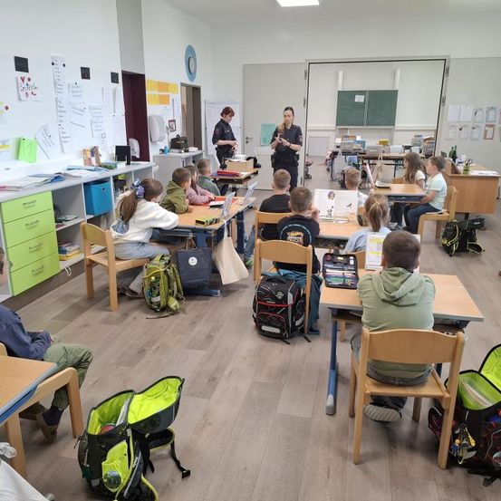 Ein Klassenzimmer mit Schülern an Schreibtischen, eine Lehrerin hält ein Buch und eine Polizistin steht an der Tür. Die Schreibtische sind in Reihen angeordnet, Schüler beschäftigen sich mit Aktivitäten.
