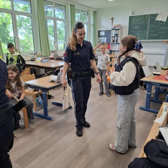 Eine Polizistin spricht mit einem Mädchen in einem Klassenzimmer mit Tischen, einer Tafel und einem Kruzifix. Andere Kinder stehen in der Nähe.