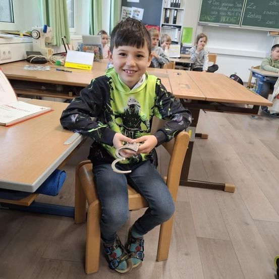 Ein junger Junge sitzt im Klassenzimmer und hält ein Paar Handschellen. Hinter ihm sitzen mehrere Kinder an Schreibtischen.
