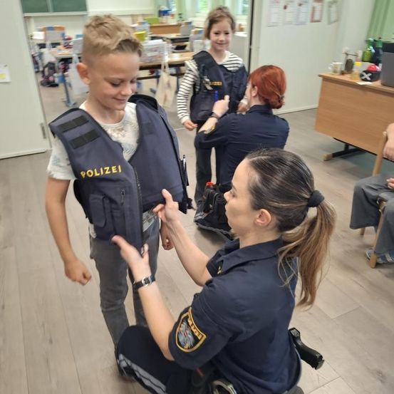 Eine Polizistin hilft einem jungen Jungen, eine blaue Weste mit der Aufschrift 'POLIZEI' anzulegen. In der Nähe steht ein Mädchen in einer ähnlichen Weste mit einer weiteren Polizistin. Eine Frau kniet und hilft dem Jungen.