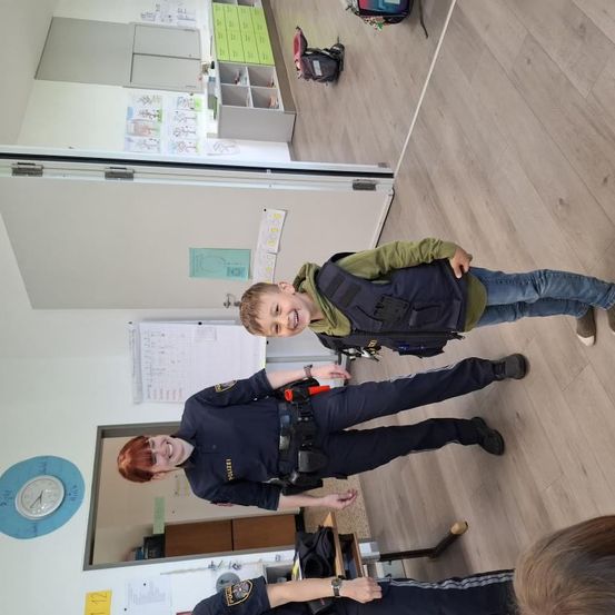 Ein lächelnder Junge mit Rucksack steht mit einem Polizisten in einem Klassenzimmer. Der Polizist trägt eine Uhr und hält eine Schlagstock. Eine Uhr ist an der Wand und ein Whiteboard ist in der Nähe.
