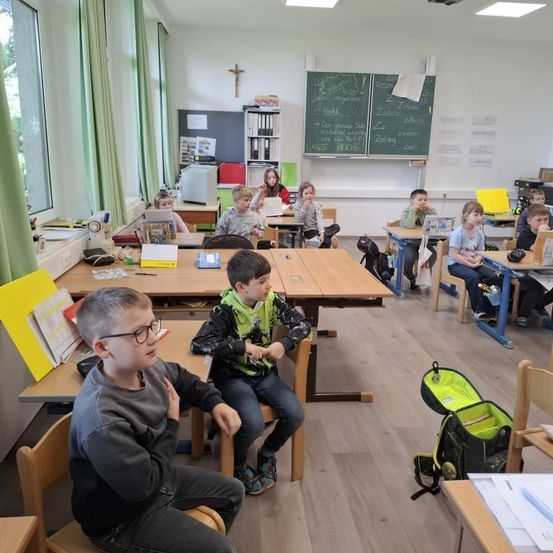 Ein Klassenzimmer mit mehreren Kindern, die an ihren Tischen sitzen, einige konzentrieren sich auf ihre Aufgaben, während andere interagieren. Eine Lehrerin steht vorne im Raum, in der Nähe einer Tafel mit Schrift darauf.
