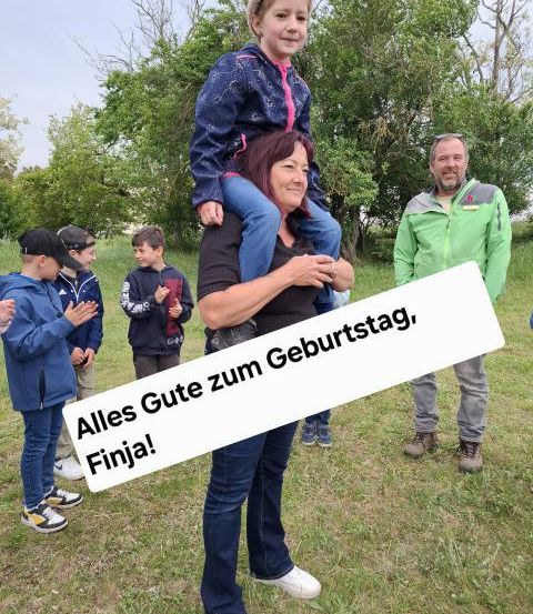 Eine Gruppe von Menschen versammelt sich in einem Park, eine Frau trägt ein Kind auf ihren Schultern. Ein Schild sagt 'Alles Gute zum Geburtstag, Finja!' Andere Kinder klatschen und lächeln.