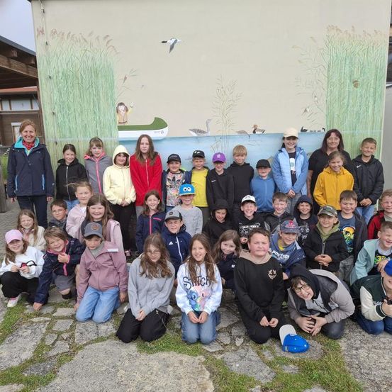 Eine Gruppe von Kindern und zwei Erwachsenen posiert für ein Foto vor einer Wandmalerei, die eine Flussszene mit Vögeln und einem Boot darstellt.