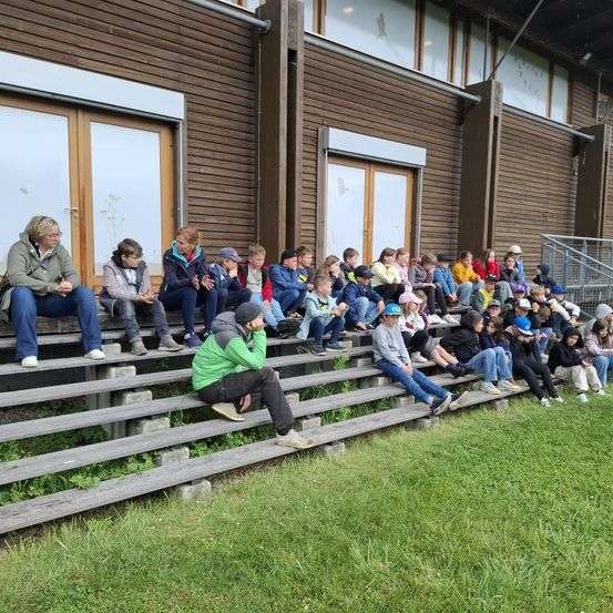 Eine Gruppe von Menschen, darunter Erwachsene und Kinder, sitzt auf den Tribünen einer Sportanlage. Einige von ihnen tragen Hüte, Brillen und Turnschuhe. Sie schauen auf etwas vor sich. Die Tribünen sind aus Holz, und der Boden ist mit Gras bedeckt.