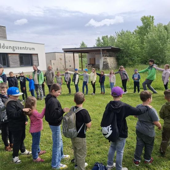 Eine Gruppe von Kindern und Erwachsenen steht auf einer Wiese. Einige tragen Rucksäcke und Turnschuhe. Dahinter befindet sich ein Gebäude mit Glasfenstern und einer Tafel mit der Aufschrift 'dungszentrum'. In der Ferne gibt es Bäume und eine Hütte.