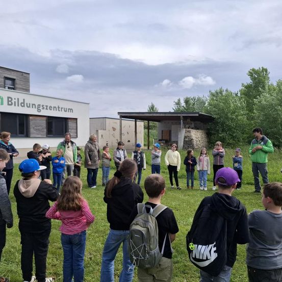 Eine Gruppe von Kindern und Erwachsenen steht auf einer Wiese. Einige tragen Hüte und Rucksäcke. Hinter ihnen steht ein Gebäude mit der Aufschrift Bildungszentrum. Im Hintergrund ein bewölkter Himmel.