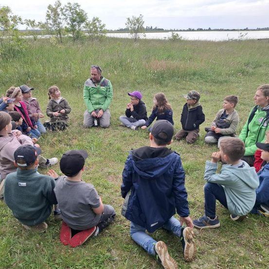 Eine Gruppe von Kindern und einem erwachsenen Mann sitzen im Kreis auf einer grasbewachsenen Fläche, möglicherweise lernen sie etwas über die Natur. Der Erwachsene trägt eine grüne Jacke. Die Kinder tragen Jacken und Mützen. Ein See ist im Hintergrund.