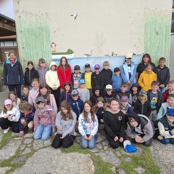 Eine Gruppe von Kindern und zwei Frauen posiert für ein Foto vor einer Wand mit einem Wandbild. Das Wandbild zeigt ein Kanu mit einer Person darin, umgeben von hohem Gras und Vögeln.