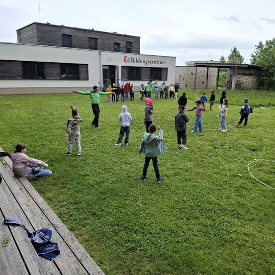 Kinder spielen auf einem Rasenfeld vor einem Gebäude mit der Aufschrift Bildungszentrum. Einige stehen, während andere auf einer Holzbank sitzen.