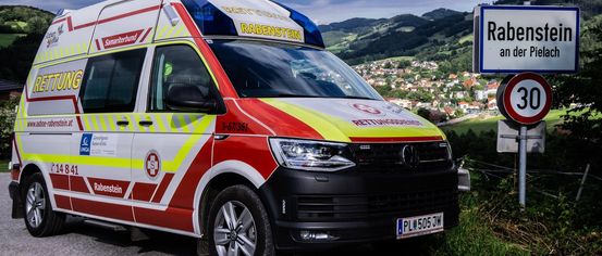 Ein Krankenwagen mit dem Kennzeichen PL 505 JW ist auf einer Straße geparkt und zeigt den Text Rettungsdienst Rabenstein. Das Fahrzeug hat ein rot-gelbes Farbschema und ist vor einem Schild geparkt. In der Ferne befinden sich Häuser und Gebäude mit Bergen im Hintergrund.