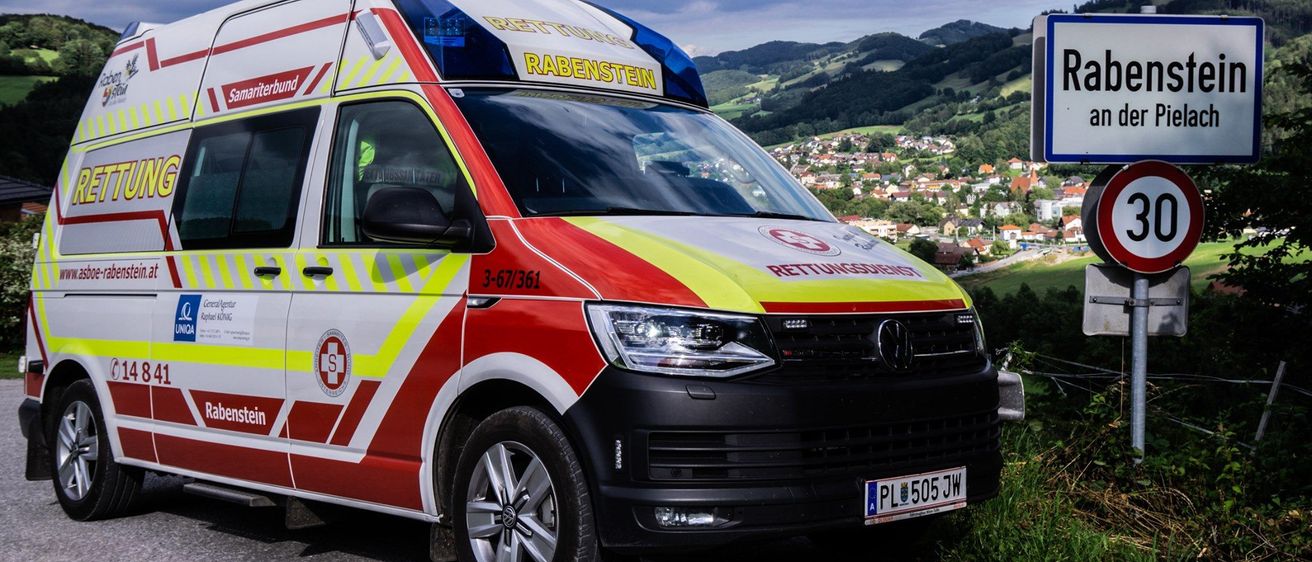 Ein Krankenwagen mit dem Kennzeichen PL 505 JW ist auf einer Straße geparkt und zeigt den Text Rettungsdienst Rabenstein. Das Fahrzeug hat ein rot-gelbes Farbschema und ist vor einem Schild geparkt. In der Ferne befinden sich Häuser und Gebäude mit Bergen im Hintergrund.