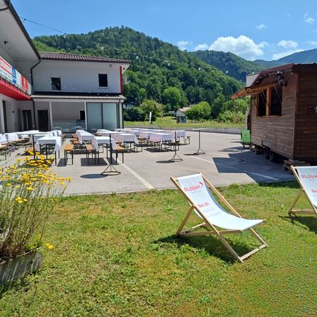 Ein Essbereich mit vielen Tischen und Stühlen vor einem Gebäude mit Bergblick. Ein hölzerner Imbisswagen ist seitlich geparkt.