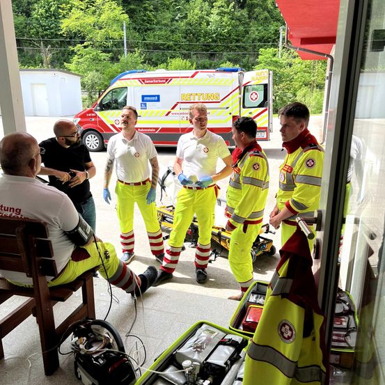Eine Gruppe von Menschen in gelben Uniformen steht in einem Gebäude, wahrscheinlich Sanitäter. Einer sitzt auf einem Stuhl, während andere in der Nähe eines Krankenwagens stehen.