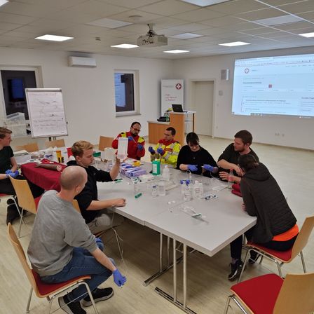 Eine Gruppe von Menschen in einem Klassenzimmer trägt Handschuhe und führt medizinische Arbeiten durch. Sie sitzen um einen Tisch mit Flaschen und anderen Gegenständen herum. An der Wand befinden sich eine Projektionsleinwand und eine Tafel.