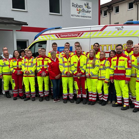 Eine Gruppe von Rettungssanitätern in leuchtend gelben Uniformen steht vor einem Krankenwagen mit der Aufschrift Samaritanbund. Hinter ihnen befindet sich ein Gebäude mit einem Schild, das Raben-Steiner Rettung Rabenstein lautet.