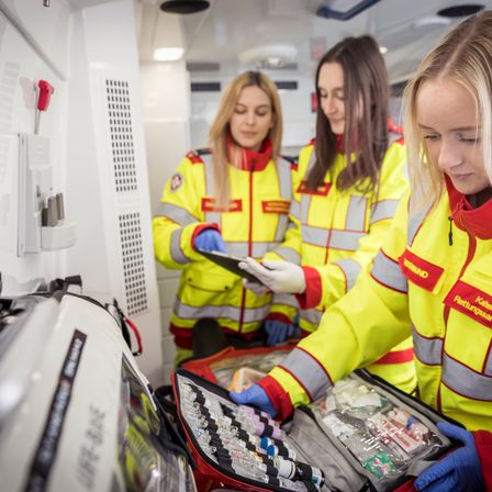 Drei Frauen in gelben Uniformen und Handschuhen, wahrscheinlich Sanitäterinnen, sind in einem Fahrzeug und überprüfen Ausrüstung. Eine hält ein Tablet, während eine andere die Inhalte einer geöffneten medizinischen Tasche betrachtet.