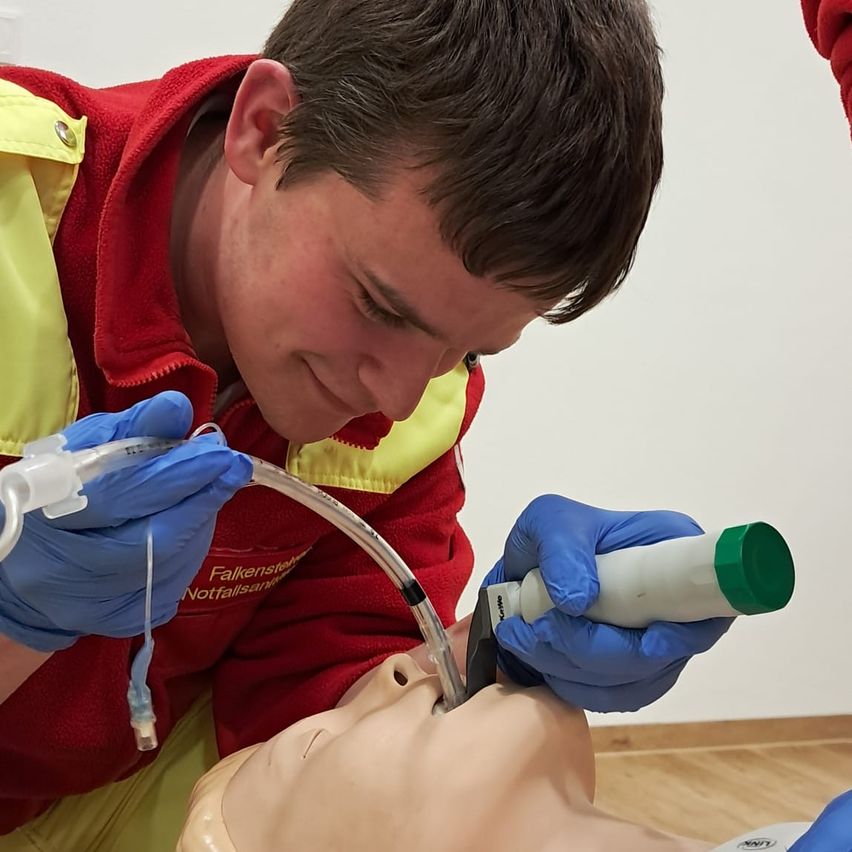 Ein junger Mann in roter und gelber Schutzkleidung übt die Anwendung eines Schlauchs an einem Mannequin. Er trägt blaue Handschuhe und scheint konzentriert auf seine Aufgabe.
