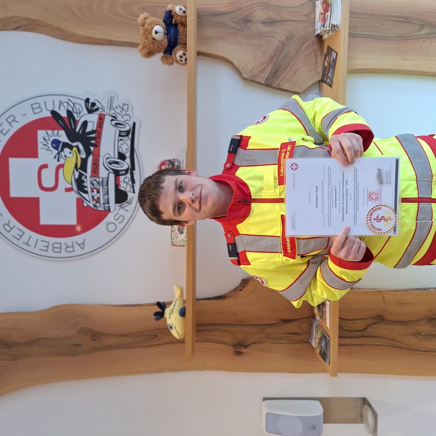 Ein junger Mann steht vor einer Wand und hält ein Zertifikat. Er trägt eine gelbe und graue Sicherheitsjacke. Hinter ihm steht ein rotes Logo mit weißem Text, das 'HELICOPTER AMBULANCE SERVICE' besagt.