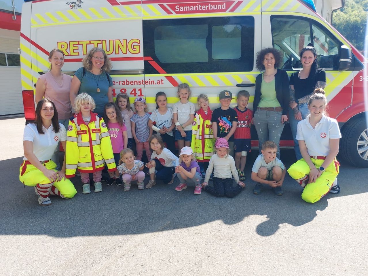 Eine Gruppe von Kindern und Erwachsenen in Warnwesten posiert für ein Foto vor einem Krankenwagen.