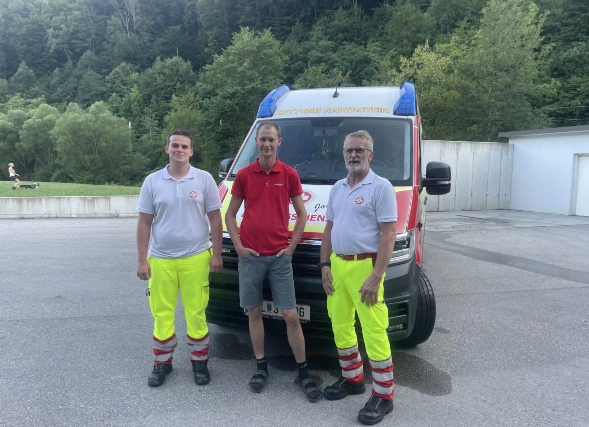Drei Männer in reflektierenden Kleidungsstücken stehen vor einem Krankenwagen mit dem Wort 'Jochen' darauf. Sie sind draußen in der Nähe einer Betonmauer mit Bäumen im Hintergrund.