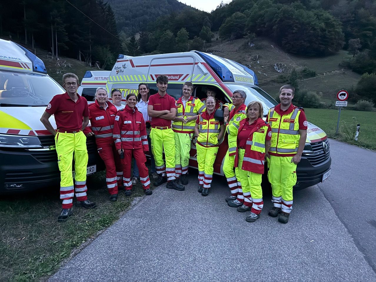 Eine Gruppe von Rettungskräften in roten und gelben Uniformen steht vor einem Krankenwagen auf einer Straße mit bergigem Hintergrund.