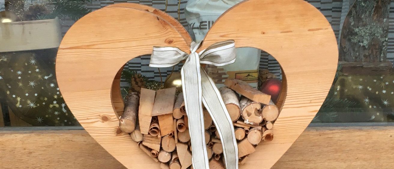Eine herzförmige Holzstruktur, gefüllt mit ordentlich gestapelten Holzscheiten und einer um sie herum gebundenen Schleife, die auf einem Holzsockel auf einem gefliesten Boden steht.