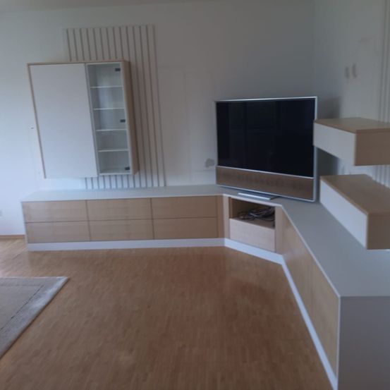 Ein modernes Wohnzimmer mit einem großen Flachbildfernseher, weißen Schränken, Holzböden und einem beigen Teppich. Der Raum hat weiße Wände und moderne Möbel.