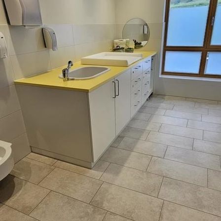 Ein Badezimmer mit gefliestem Boden und einer Arbeitsplatte mit zwei Waschbecken, einem Spiegel und Schränken. Der Raum hat auch eine Toilette, einen Handtrockner und eine Papiertuchspender.