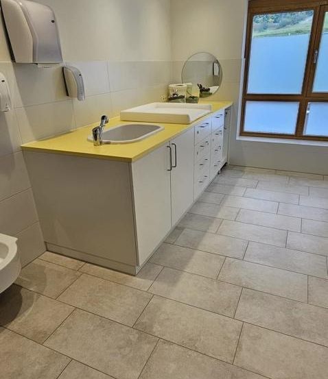 Ein Badezimmer mit gefliestem Boden und einer Arbeitsplatte mit zwei Waschbecken, einem Spiegel und Schränken. Der Raum hat auch eine Toilette, einen Handtrockner und eine Papiertuchspender.