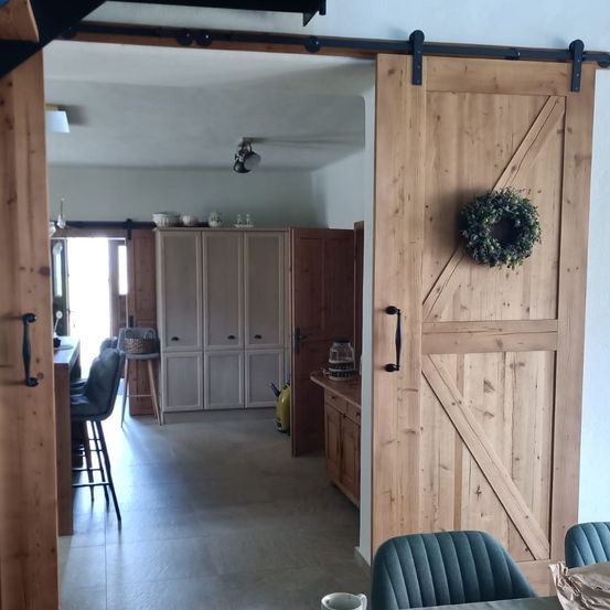 Eine offene Schiebetür führt in eine moderne Küche. Die Küche hat Holzschränke und eine Arbeitsplatte mit Gegenständen darauf. An der Tür hängt ein Kranz, und ein Essbereich ist mit Stühlen und einem Tisch zu sehen.