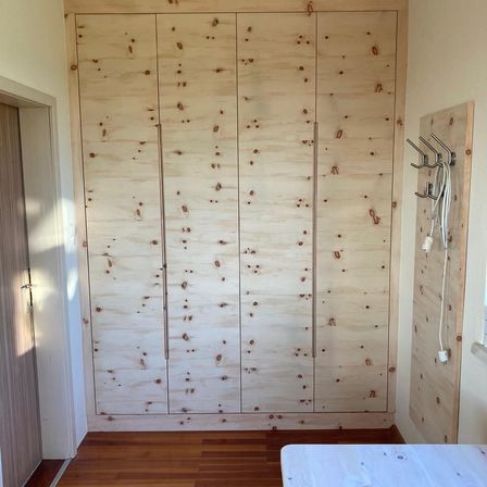 Ein Innenraum einer Zimmer mit einem großen Holzschrank an der Wand. Der Schrank hat ein strukturiertes Kieferndesign mit sichtbaren Ästen. Rechts befindet sich ein Holzpegboard mit einigen hängenden Kabeln. Der Boden ist aus Holz und ein weißes Bett steht auf der rechten Seite.