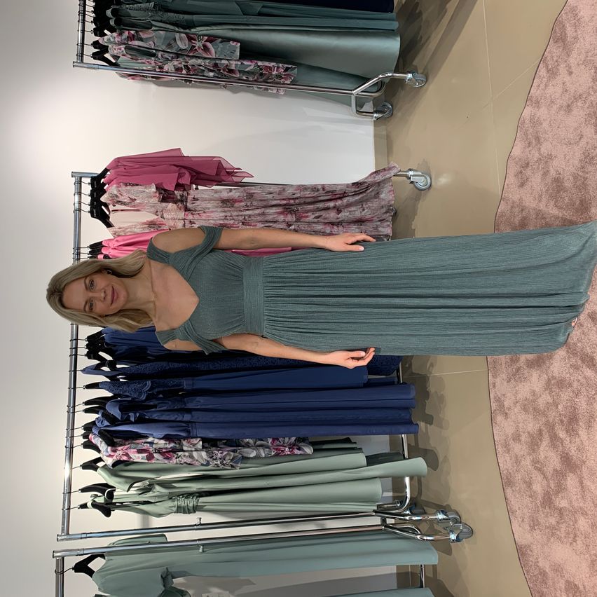 Eine Frau in einem grauen Kleid steht zwischen Kleiderständern in einem Bekleidungsgeschäft.