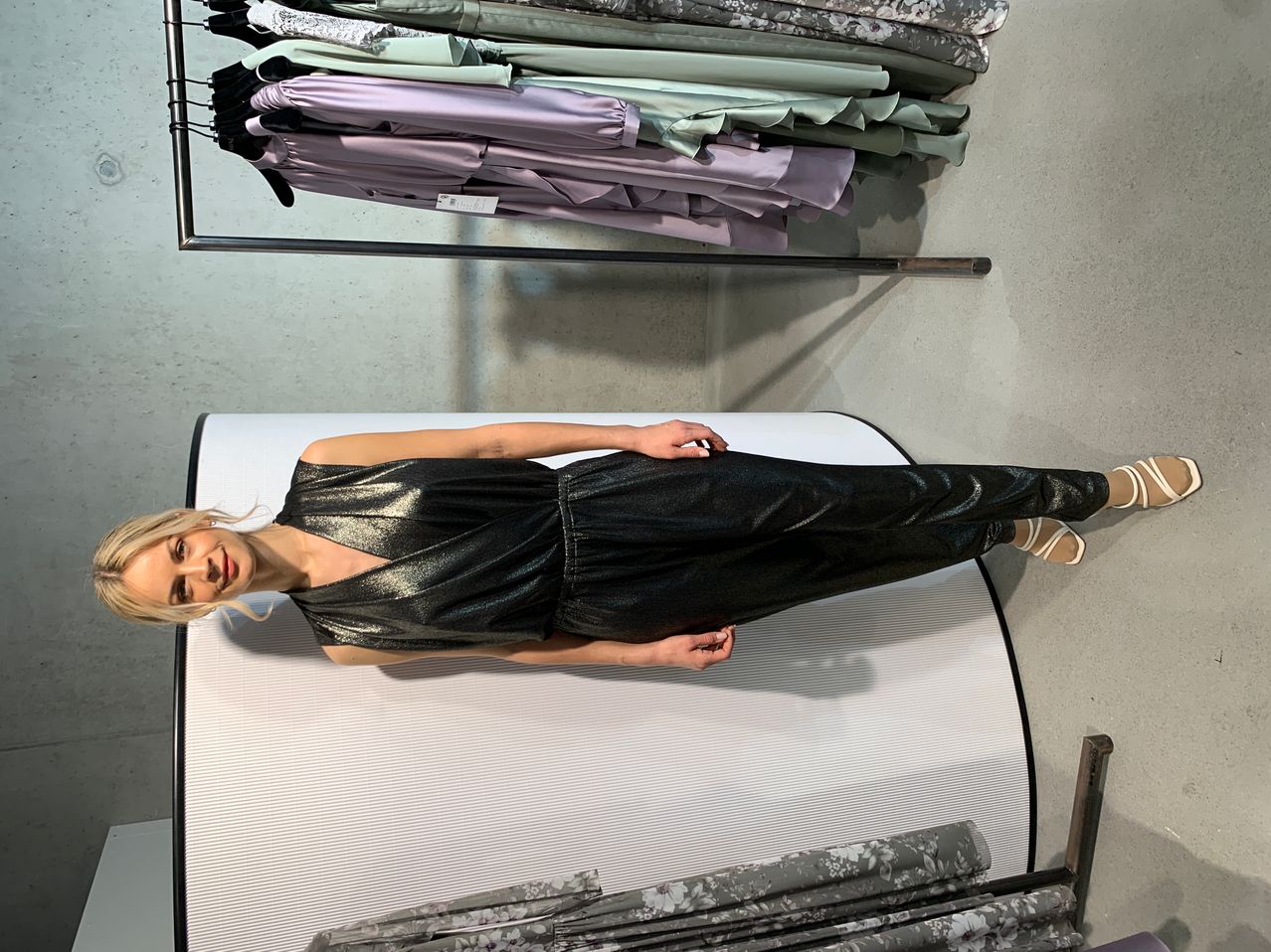 A woman in a black metallic dress poses in front of a clothing rack with folded dresses in various colors.