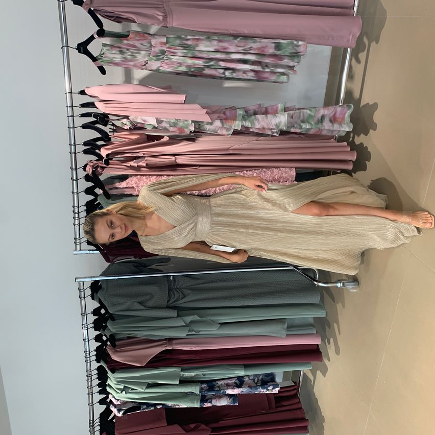 A blonde woman lies on a rack of dresses. The dresses are in various colors, including pink, beige, and green.