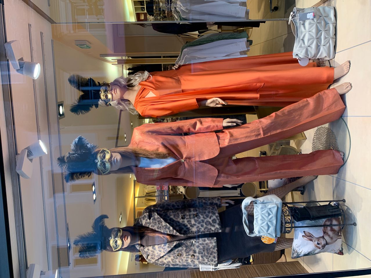 Vier Mannequins in Masken und schicken Outfits sind in einem Schaufenster ausgestellt. Die Mannequins tragen orangefarbene Kleider und Mäntel.