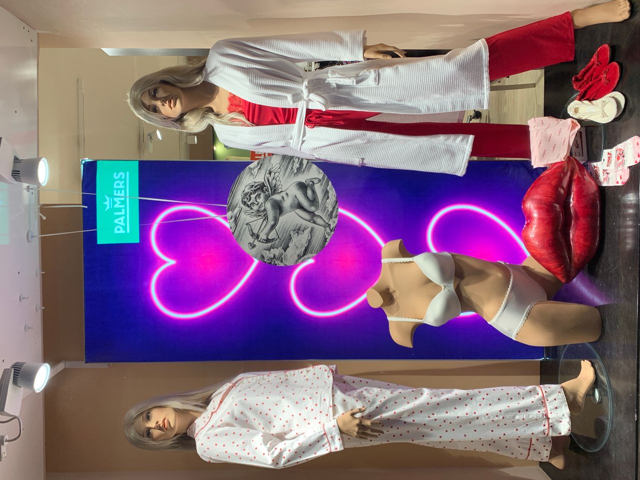 Drei Mannequins in Dessous und Schlafanzügen stehen vor einem blauen Neonschild. Das oberste Mannequin trägt einen weißen Bademantel, das mittlere einen weißen BH und Höschen und das unterste rote Dessous.