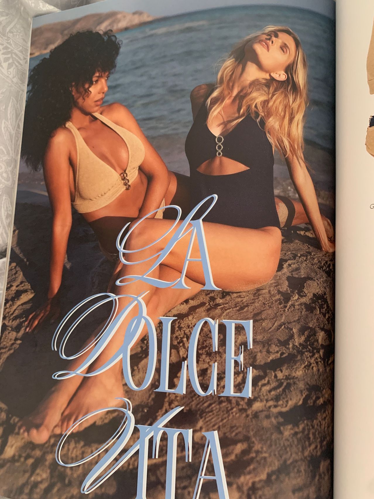 Zwei Frauen sitzen auf einem sandigen Strand und tragen Bademode. Eine hat einen beigen Bikini und die andere einen schwarzen. Das Wort 'LA DOLCE' ist in Blau auf den Sand geschrieben.