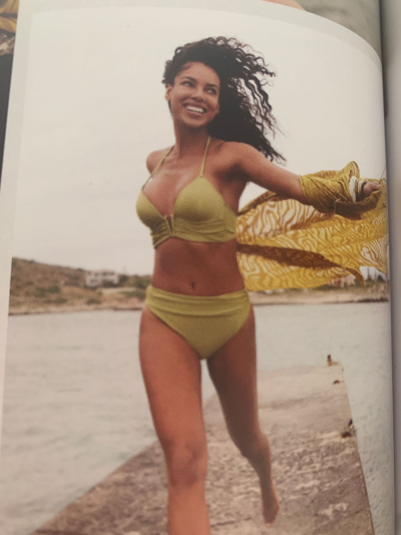 Eine Frau in einem grünen Bikini steht auf einem Pier, lächelt und posiert für ein Foto. Der Hintergrund zeigt einen ruhigen Strand mit ruhigem Wasser.