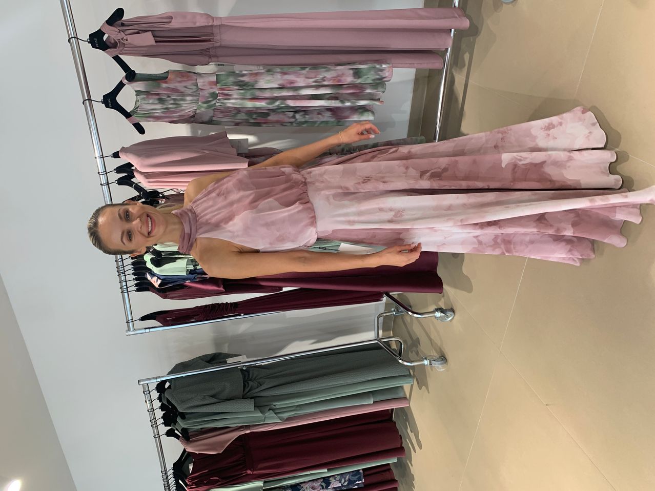 Eine Frau in einem rosa Kleid steht vor einem Kleiderständer mit Kleidern in verschiedenen Farben, die an Kleiderbügeln hängen.