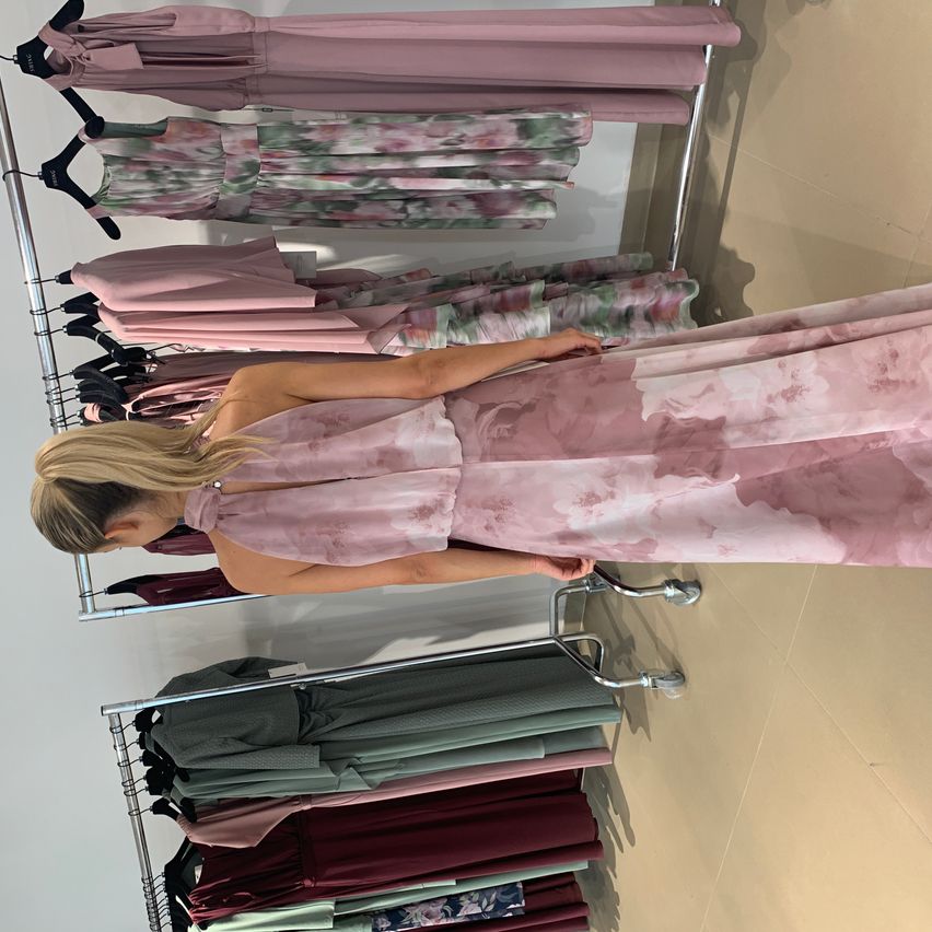 Eine Frau betrachtet ein rosa Blumenkleid in einem Bekleidungsgeschäft. Das Kleid ist auf einem Ständer ausgestellt, zusammen mit anderen Kleidern in verschiedenen Farben und Stilen.