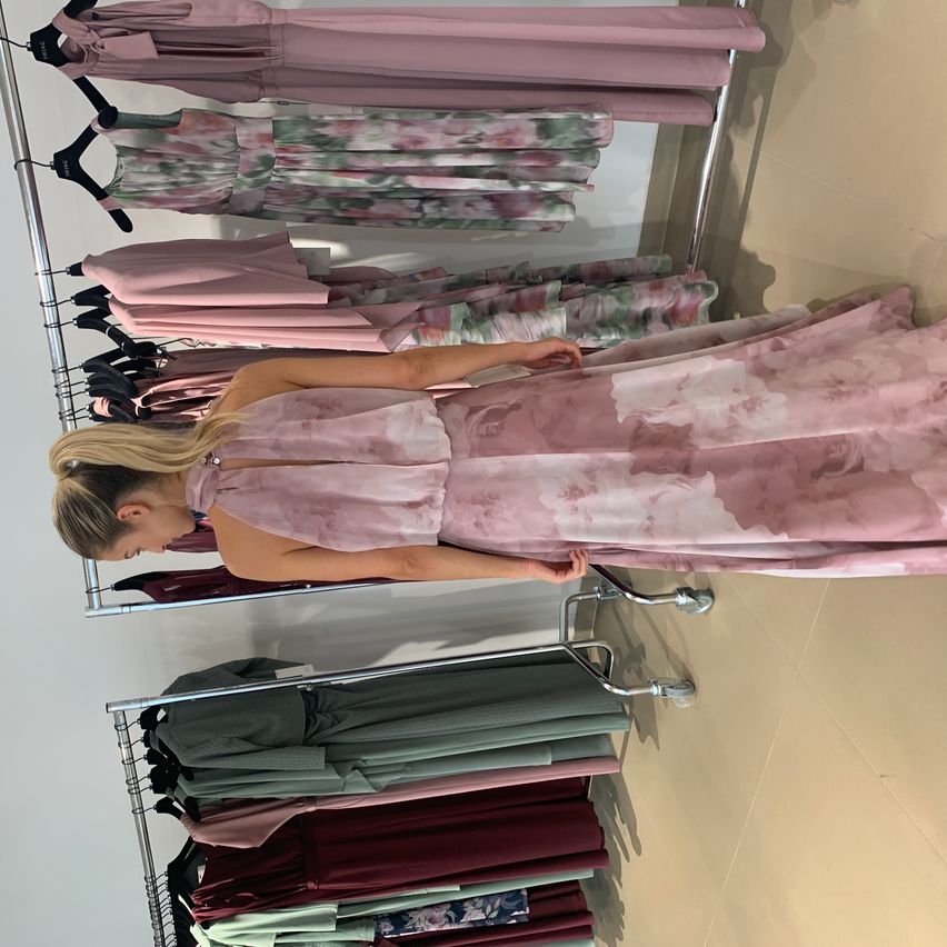 Eine Frau in einem rosa Blumenkleid steht vor einem Kleiderständer mit Kleidern an Kleiderbügeln, einschließlich rosa Blumen- und Weinrotkleidern.