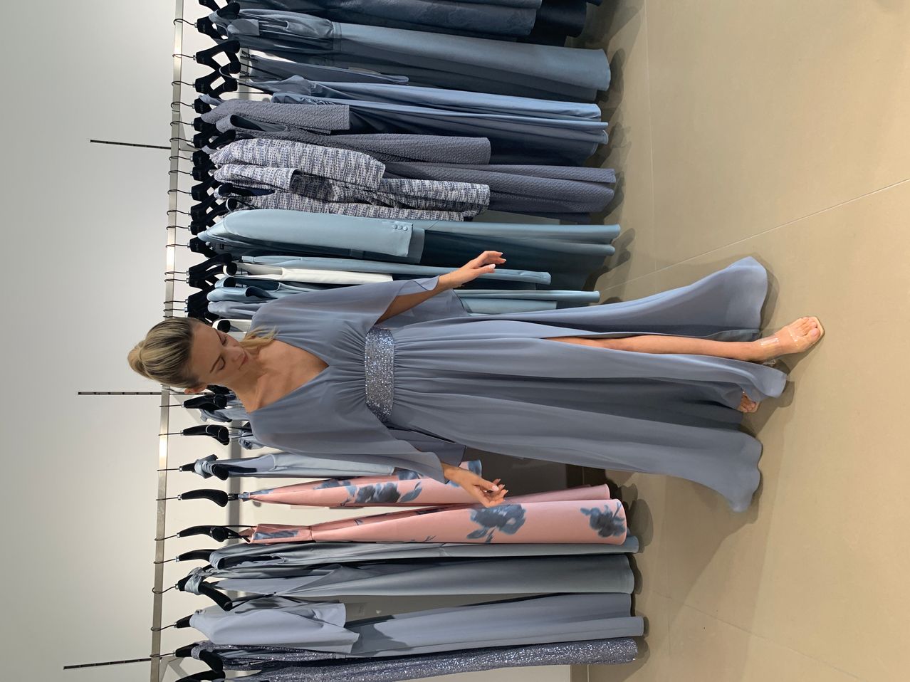 Eine Frau posiert vor einer Kulisse aus ordentlich gefalteten Kleidern. Sie trägt ein blaues Kleid und blickt nach unten, eine Hand auf einem Kleid ruhend.