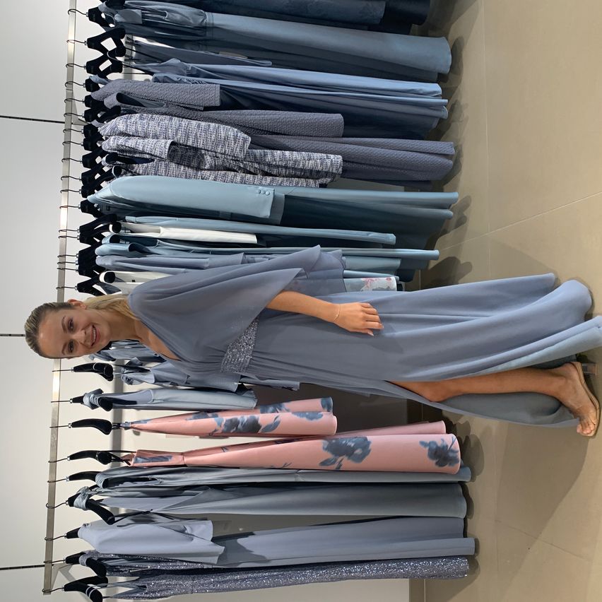 Eine Frau in einem blauen Kleid steht vor einem Kleiderständer und lächelt. Die Kleider sind überwiegend blau und rosa.