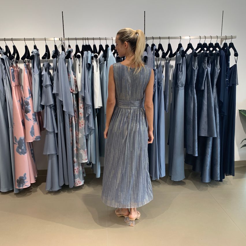Eine Frau in einem blauen Kleid steht vor einem Kleiderständer mit Kleidern in verschiedenen Farben und Stilen.