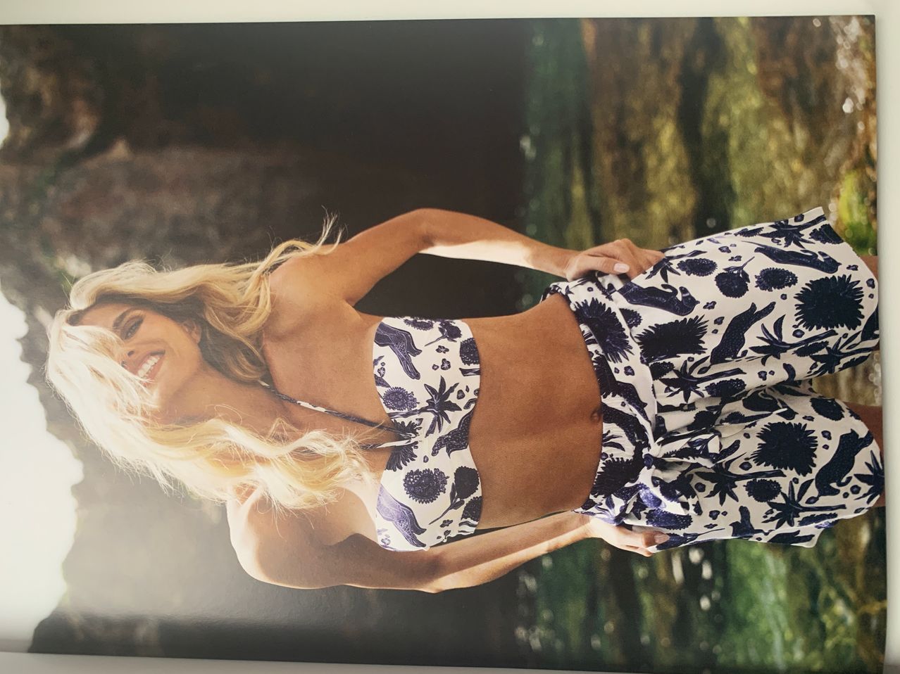 Eine Frau mit blonden Haaren lächelt in die Kamera und trägt einen weiß-blau gemusterten Bikini. Sie liegt auf einem grünen Hintergrund.