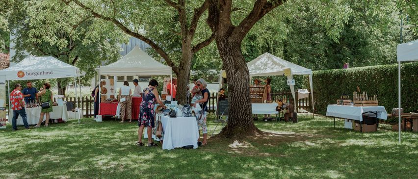 Eine Gruppe von Menschen, die an einem sonnigen Tag unter Zelten nach Artikeln suchen, einige stehen, andere sitzen, mit Bäumen und einem Holzzaun im Hintergrund.